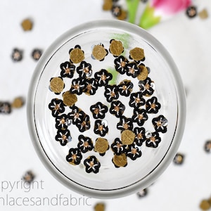 May include: A close-up view of a glass bowl filled with black and gold flower-shaped sequins. The sequins are scattered on a white surface.