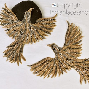 May include: Two gold-coloured bird appliques with beaded details. The birds are in a flying pose with their wings spread out.