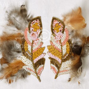 Puede incluir: Dos apliques de plumas con cuentas de color rosa, blanco, dorado y marrón. Los apliques están rodeados de plumas marrones y grises.