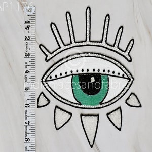 May include: A white and green evil eye patch with black and white details. The eye is surrounded by white sequins and has a green iris. The patch is approximately 12.7 cm in length.