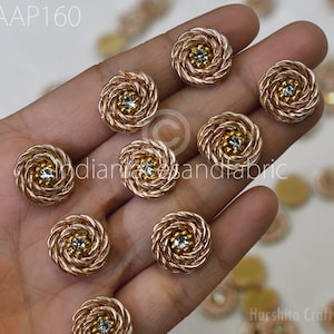 Pode incluir: Um close-up de dez botões decorativos redondos dourados com um design em espiral e um strass transparente no centro. Os botões são segurados em uma mão.