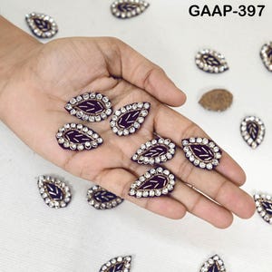 以下が含まれることがあります： 葉の形をした装飾品のコレクション。各ピースは、濃い紫色の中心部、金色のステッチ、透明なラインストーンの縁取りが特徴です。「GAAP-397」の文字が見えます。