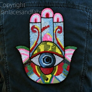 以下が含まれることがあります： 濃い青色のデニムジャケット。背面には、カラフルなビーズで装飾された、眼のデザインが施されたハンズオブファティマが描かれています。