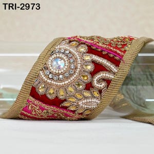Pode incluir: Close-up de uma guarnição decorativa com base de veludo vermelho, adornada com contas douradas, brancas e transparentes. A guarnição tem uma borda dourada e detalhes em rosa. O texto "TRI-2973" é visível.
