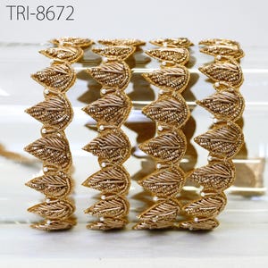 May include: Four gold-toned bangles featuring a repeating leaf-shaped pattern with metallic thread embroidery and small white bead accents. The text TRI-8672 is visible in the upper left corner of the image. The bangles have a textured finish.