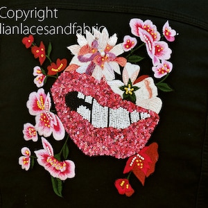 以下が含まれることがあります： ピンクと白の刺繍の花柄とピンクと白のビーズでできた唇のデザインが施された黒いデニムジャケット。ジャケットには「Copyright Indianlacesandfabric」という文字が刺繍されている。