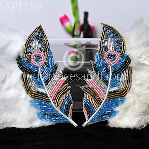 May include: Two blue, gold, and pink beaded feather appliques with white feathers on either side. The appliques are on a clear glass surface.