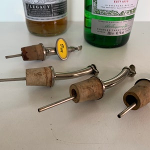 May include: Four silver bottle pourers with cork stoppers. The pourers have a long, curved spout and a small, round handle. One of the pourers has a yellow tag that says "Tart".