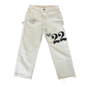 May include: Off-white denim jeans with a straight leg and frayed hem. The jeans feature a black graphic of a smiling face with text on the left leg and the number "22" on the right leg. The jeans have a classic five-pocket design.