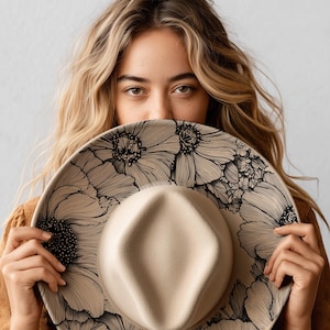 Puede incluir: Sombrero fedora beige con ala ancha, decorado con un dise&ntilde;o floral negro. La copa del sombrero es de color beige claro. El ala est&aacute; adornada con grandes y detalladas ilustraciones florales negras. El sombrero se sostiene frente a una persona.