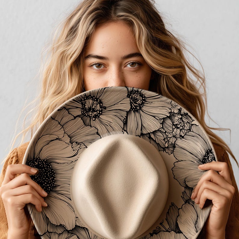 Puede incluir: Sombrero fedora beige con ala ancha, decorado con un dise&ntilde;o floral negro. La copa del sombrero es de color beige claro. El ala est&aacute; adornada con grandes y detalladas ilustraciones florales negras. El sombrero se sostiene frente a una persona.