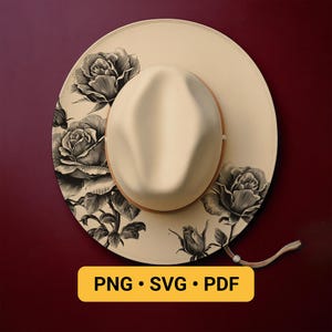 Op de afbeelding: Een crèmekleurige hoed in fedora-stijl met een brede rand, versierd met zwart-witte rozenillustraties. De hoed heeft een bruine band rond de kroon en een kinriem. De onderkant van de afbeelding heeft een gele rechthoek met de tekst "PNG • SVG • PDF".