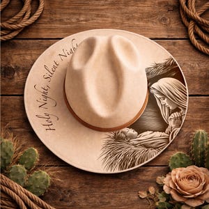 May include: A beige felt hat with a brown leather band featuring a dark brown illustration of a nativity scene and the text "Holy Night, Silent Night" printed in a cursive script on the wide brim of the hat, viewed from directly above.