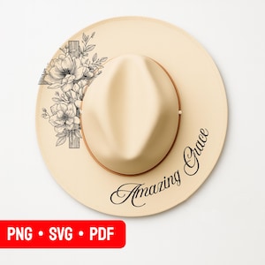 May include: A cream-colored fedora hat with a brown band and a floral cross design. The hat features the words "Amazing Grace" in a script font. The hat is a stylish accessory.