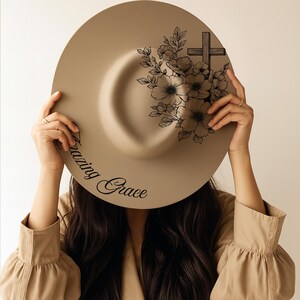 May include: A beige fedora hat with a flat brim. The hat has a black floral and cross design on the crown and the words "Amazing Grace" in script. The hat is held up by a person.