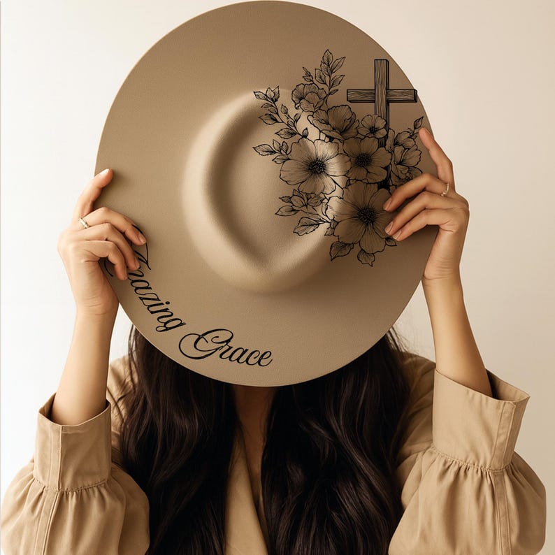 May include: A beige fedora hat with a flat brim. The hat has a black floral and cross design on the crown and the words "Amazing Grace" in script. The hat is held up by a person.