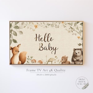 May include: A framed print with the text "Hello Baby" in a whimsical font. The artwork features woodland creatures like a fox, bear, and hedgehog, along with mushrooms and foliage in soft, natural tones. The frame is a light wood color.