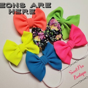 May include: A collection of colorful hair bows in neon shades of pink, green, yellow, and blue, with one bow featuring a floral pattern. The bows are attached to white headbands. The text "NEONS ARE HERE" is displayed at the top.