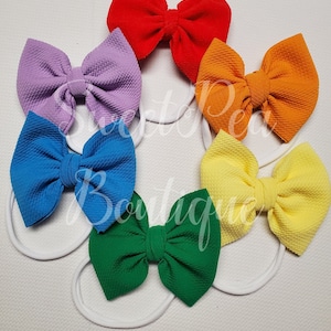 May include: Six colorful hair bows with white elastic bands. The bows are made of a textured fabric and are arranged in a rainbow pattern. The colors are red, purple, orange, blue, yellow, and green.