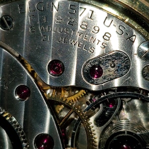 May include: Close-up of a silver watch movement with the text "ELGIN 571U-SA F 192898 8 ADJUSTMENTS 21 JEWELS" visible.