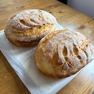 May include: Two round loaves of freshly baked bread with golden-brown crusts and decorative cuts on top. The bread sits on a sheet of white parchment paper, ready to be sliced. The loaves are on a wooden surface.