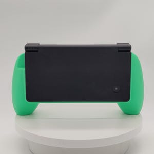 以下が含まれることがあります： マットブラックの長方形の本体に、鮮やかなミントグリーンのシリコン製グリップカバーを装着した携帯型ゲーム機。デバイスの背面は滑らかで無地であり、右下には小さな円形のカメラレンズが配置されています。