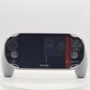 Pode incluir: Uma consola de jogos portátil Sony PS Vita com ecrã preto, moldura prateada e pegas de comando cinzentas integradas em ambos os lados. O dispositivo apresenta o logótipo da Sony no canto superior esquerdo e o logótipo PS Vita ao centro.