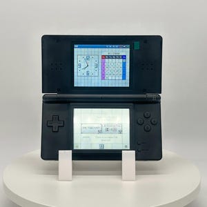 May include: A black Nintendo DS handheld game console, open to display two screens. The top screen shows a clock and calendar, while the bottom screen displays text. The console is resting on a white stand.