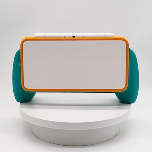 May include: A handheld gaming console grip featuring a white textured back panel, a vibrant orange rectangular frame, and teal ergonomic side handles. The device has a white top edge with a central black circular port and a smooth, clean design.