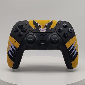 Peut inclure: Une manette de jeu vidéo sans fil noire avec un motif graphique jaune et noir. Le pavé tactile central affiche une illustration stylisée d'un personnage masqué aux dents découvertes, tandis que les poignées présentent des griffures jaunes.