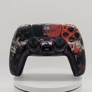 May include: A wireless video game controller featuring a custom black and red graphic wrap. The design includes detailed illustrations of skulls and figures. The central touchpad displays Japanese text, and the analog sticks have skull icons.
