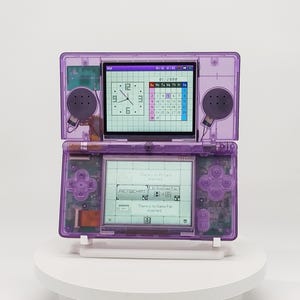 Peut inclure: Une console de jeu portable violette translucide avec un design à clapet à double écran. L'écran supérieur affiche une horloge et un calendrier pour janvier 2008. L'écran inférieur montre un menu avec le texte PictChat et un message d'erreur.