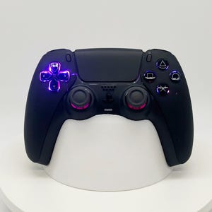 Op de afbeelding: Een matzwarte draadloze videogamecontroller met geïntegreerde paarse LED-verlichting achter de richtingsknop en actieknoppen. De controller bevat twee analoge sticks met paarse ringaccenten en een centraal aanraakgevoelig paneel.