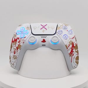 Op de afbeelding: Een witte draadloze gamecontroller met een aangepast grafisch ontwerp met gouden schedel- en roosillustraties, rode verfspatten en een roze X-symbool op het centrale touchpad. De controller heeft blauwe LED-lichtringen rond de joysticks.