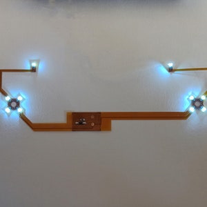 Może przedstawiać: Obwód elektroniczny z czterema świecącymi na niebiesko światłami ułożonymi w kształt krzyża, połączonymi cienką, pomarańczowo-brązową elastyczną płytką obwodu. Dwa dodatkowe niebieskie światła na końcach.