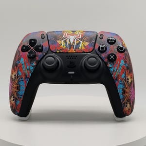 May include: A black video game controller featuring a vibrant, multi-colored paint splatter design across the front shell. A white spider emblem is centered on the touchpad, surrounded by black buttons, joysticks, and a directional pad.
