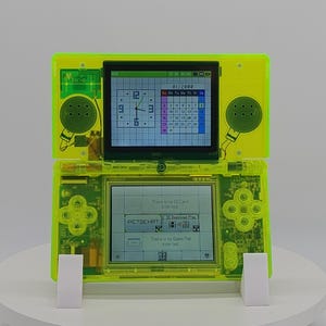 Op de afbeelding: Een felgroene handheld gameconsole met twee schermen. Het bovenste scherm toont een klok en kalender, het onderste scherm toont tekst. De console heeft een D-pad, knoppen en luidsprekers. Hij staat op een witte standaard.
