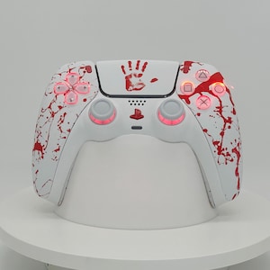 May include: A white video game controller featuring a red handprint graphic on the centre touchpad and red paint splatter patterns across the grips. The controller includes illuminated red buttons and analogue sticks with a central red logo.
