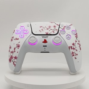 May include: A white wireless video game controller featuring a pink cherry blossom branch pattern across the front shell. The device includes a red PlayStation logo, two white analogue sticks, and buttons illuminated with a soft purple light.