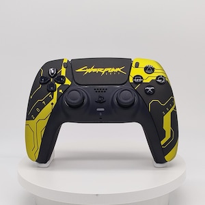 May include: A black and yellow wireless video game controller featuring the Cyberpunk 2077 logo on the central touchpad. The controller has a matte black base with yellow geometric circuit-like patterns and the number 2077 printed on the grips.