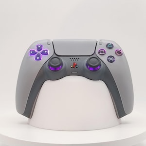 Könnte beinhalten: Ein grauer kabelloser Videospiel-Controller mit einem mehrfarbigen PlayStation-Logo in der Mitte, zwei Analogsticks mit violetten Akzenten sowie einem Steuerkreuz und Aktionstasten, die in einem leuchtenden Violett auf dem grauen Gehäuse erstrahlen.