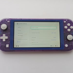 May include: A purple Nintendo Switch Lite console with a white screen displaying the system settings menu. The menu includes options for system update, themes, notifications, sleep mode, controllers and sensors, and system. The current system version is 16.0.3.