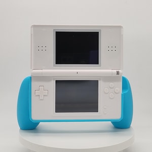Könnte beinhalten: Eine weiße tragbare Videospielkonsole mit zwei Bildschirmen, einem Steuerkreuz, Aktionstasten und einem hellblauen, ergonomischen Griffaufsatz, der die Seiten des Geräts umschließt und für eine bessere Handhabung beim Spielen sorgt.