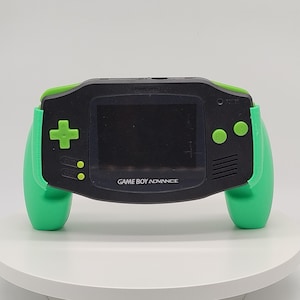 Può includere: Una console di gioco portatile Game Boy Advance nera dotata di un'impugnatura ergonomica verde brillante. Il dispositivo include uno schermo centrale, un pad direzionale, due pulsanti azione e i tasti start e select, tutti di colore verde.