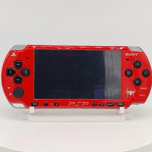 May include: A red Sony PSP handheld gaming console featuring a black screen, silver accents, and a white spider graphic on the right side. The device includes standard directional buttons, action buttons, and the PSP logo printed in white on the front.
