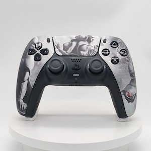 Puede incluir: Un mando de juego inalámbrico en blanco y negro con un estampado gráfico detallado de un personaje musculoso en la placa frontal. El mando incluye dos joysticks negros, una cruceta, cuatro botones de acción y un logotipo central.