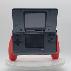 以下が含まれることがあります： ダークグレーのNintendo DS携帯ゲーム機。上部画面と下部タッチスクリーンのデュアルスクリーンデザインが特徴です。黒い十字キーとアクションボタンを備え、本体の両サイドには明るい赤色の人間工学に基づいたグリップが装着されています。
