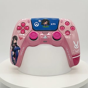 Puede incluir: Un mando de videojuegos rosa con gráficos del personaje D.Va, un icono de conejo blanco y el texto "LOVE D.VA" e "I PLAY TO WIN!". El panel táctil muestra un personaje pixelado y el logotipo de Overwatch en un área rectangular azul.