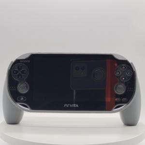 Può includere: Una console di gioco portatile Sony PS Vita grigia. La console ha uno schermo nero con pulsanti di controllo su entrambi i lati. La parola "SONY" è visibile sopra i pulsanti di controllo sinistri e "PSVita" è stampato sotto.
