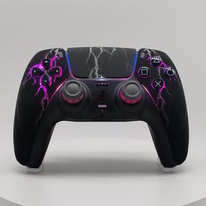 May include: A black wireless video game controller featuring a purple and white lightning bolt pattern across the front shell. The device includes two analog sticks with pink accents, a directional pad, four action buttons, and a central logo.